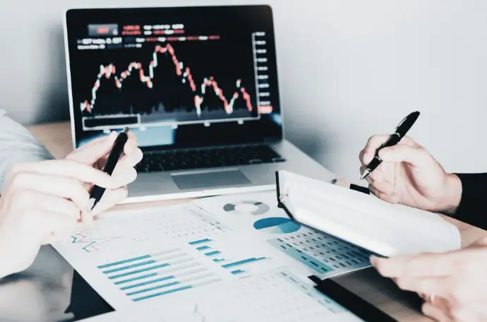 Manos de dos profesionales en reunión de asesoría mercantil, con bolígrafo y papel mientras analizan datos en un ordenador