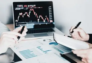 Manos de dos profesionales en reunión de asesoría mercantil, con bolígrafo y papel mientras analizan datos en un ordenador